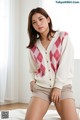 A woman sitting on a bed wearing a pink and white argyle cardigan.